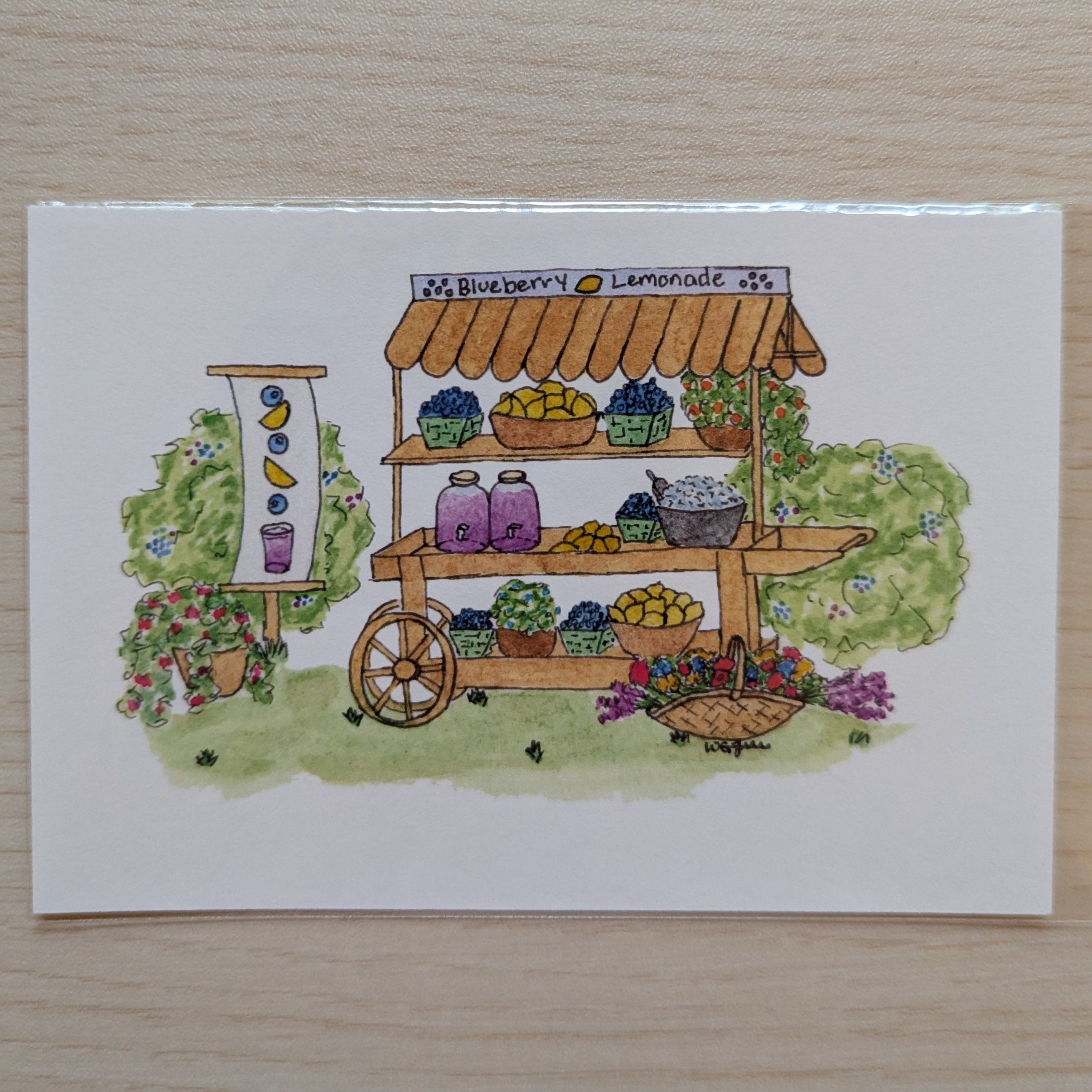 A square white art print featuring a watercolor illustration of a blueberry lemonade cart with greenery, flowers, and colorful jars, placed on a light wood surface.