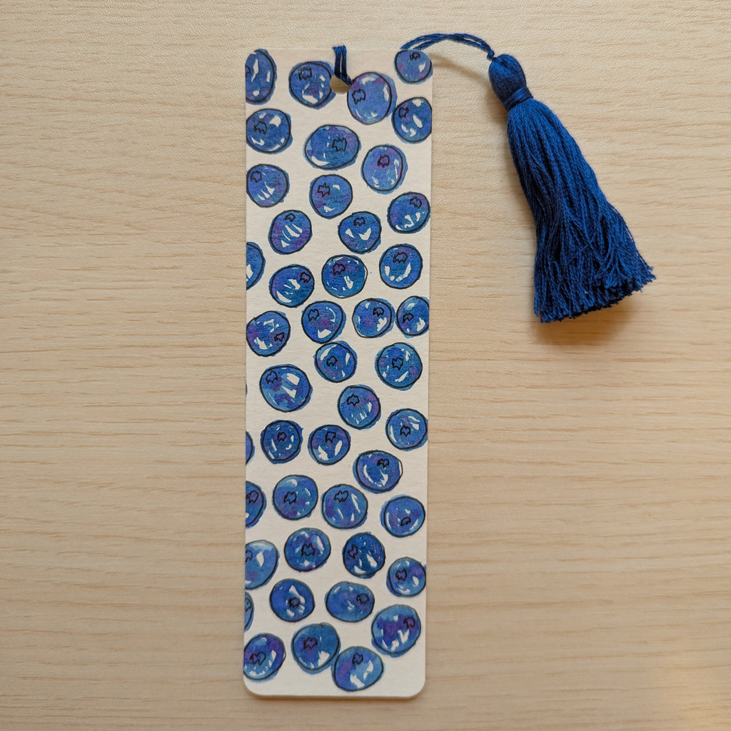 A rectangular paper bookmark with a blueberries on a white background, featuring a blue tassel attached at the top, displayed on a light wood surface.
