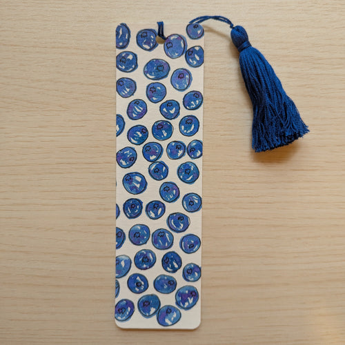 A rectangular paper bookmark with a blueberries on a white background, featuring a blue tassel attached at the top, displayed on a light wood surface.