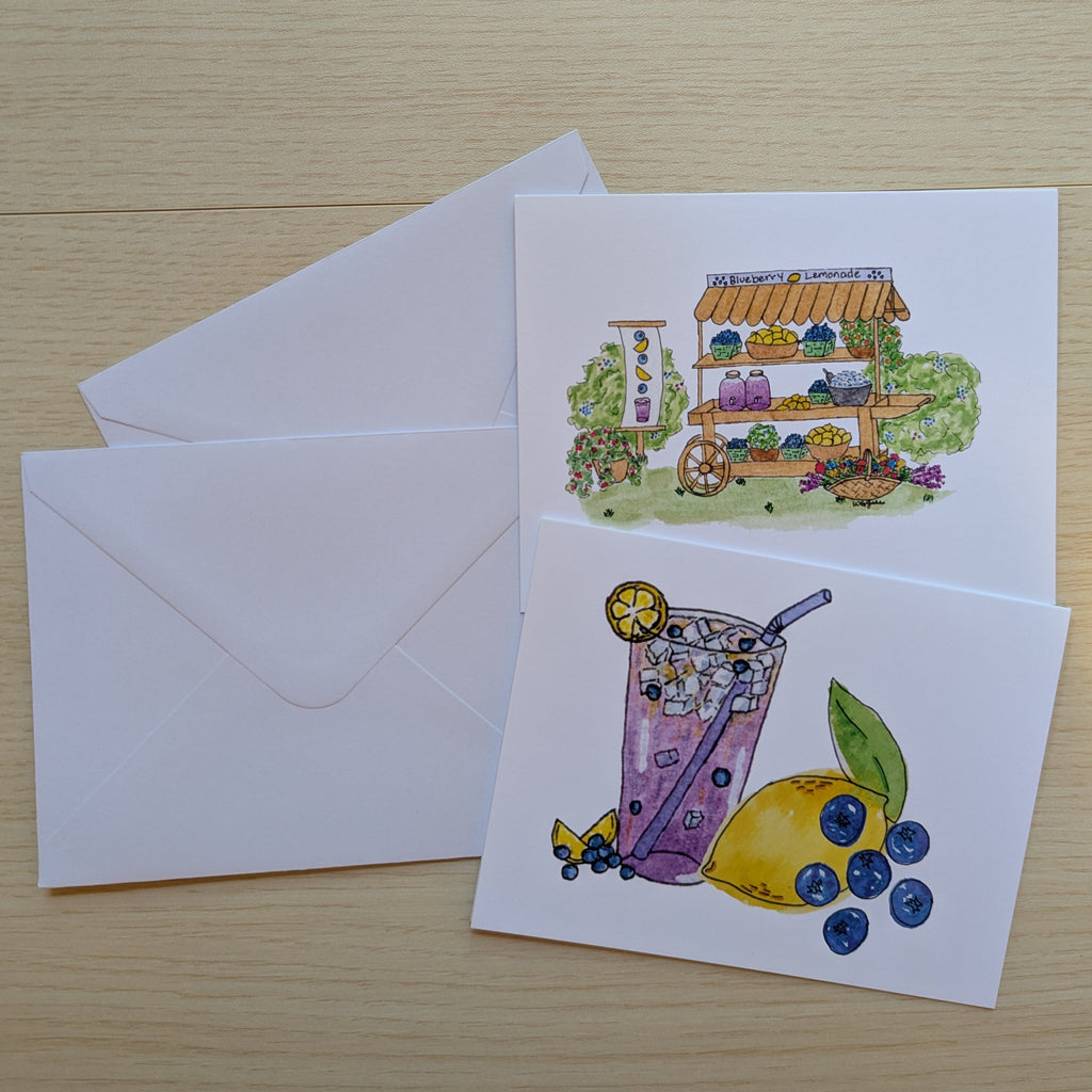 Two white illustrated greeting cards with white envelopes on a light wood surface. The front card features a  purple drink with blueberries and a lemon and the back card features colorful watercolor-style drawing of a lemonade cart.
