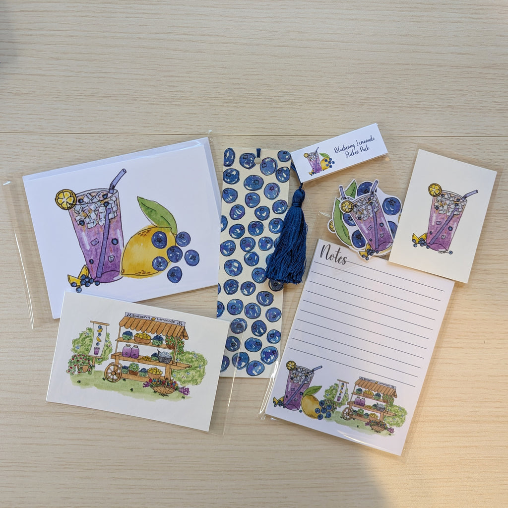 A flat lay of small stationery items on a light wood surface, including illustrated cards, a lined note pad, a hand painted blueberry bookmark with a tassel, blueberry-themed stickers, and two small illustrated art prints. The designs feature purple iced drinks, blueberries, and a lemonade stand illustration.