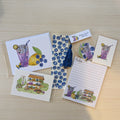 A flat lay of small stationery items on a light wood surface, including illustrated cards, a lined note pad, a hand painted blueberry bookmark with a tassel, blueberry-themed stickers, and two small illustrated art prints. The designs feature purple iced drinks, blueberries, and a lemonade stand illustration.
