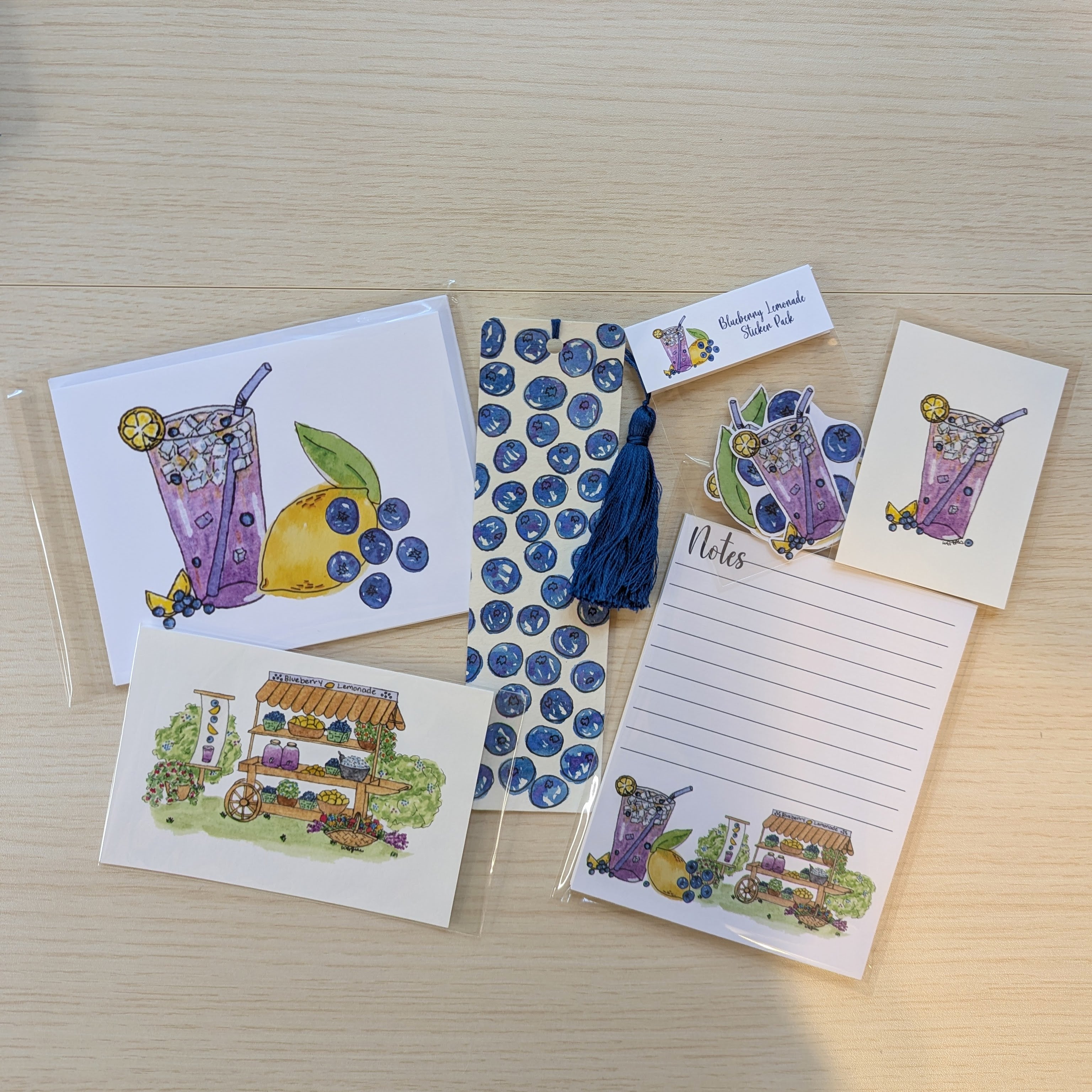 A flat lay of small stationery items on a light wood surface, including illustrated cards, a lined note pad, a hand painted blueberry bookmark with a tassel, blueberry-themed stickers, and two small illustrated art prints. The designs feature purple iced drinks, blueberries, and a lemonade stand illustration.