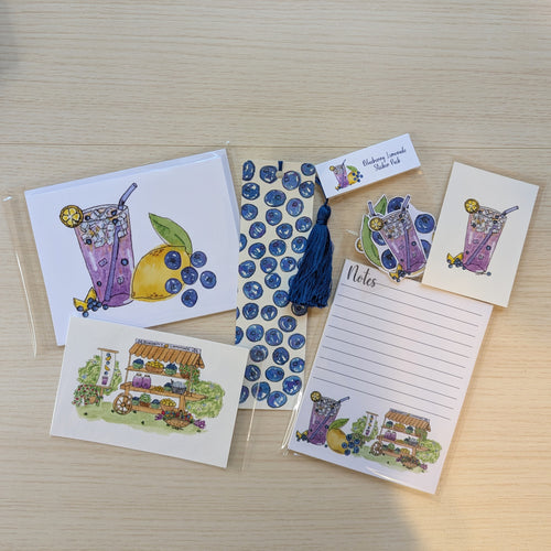 A flat lay of small stationery items on a light wood surface, including illustrated cards, a lined note pad, a hand painted blueberry bookmark with a tassel, blueberry-themed stickers, and two small illustrated art prints. The designs feature purple iced drinks, blueberries, and a lemonade stand illustration.