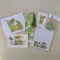 A coordinated stationery set laid out on a light wood surface, featuring illustrated kiwi slices, a drink with a straw, a small market scene, lined notes paper, a hand painted kiwi bookmark with a tassel, and a sticker set.