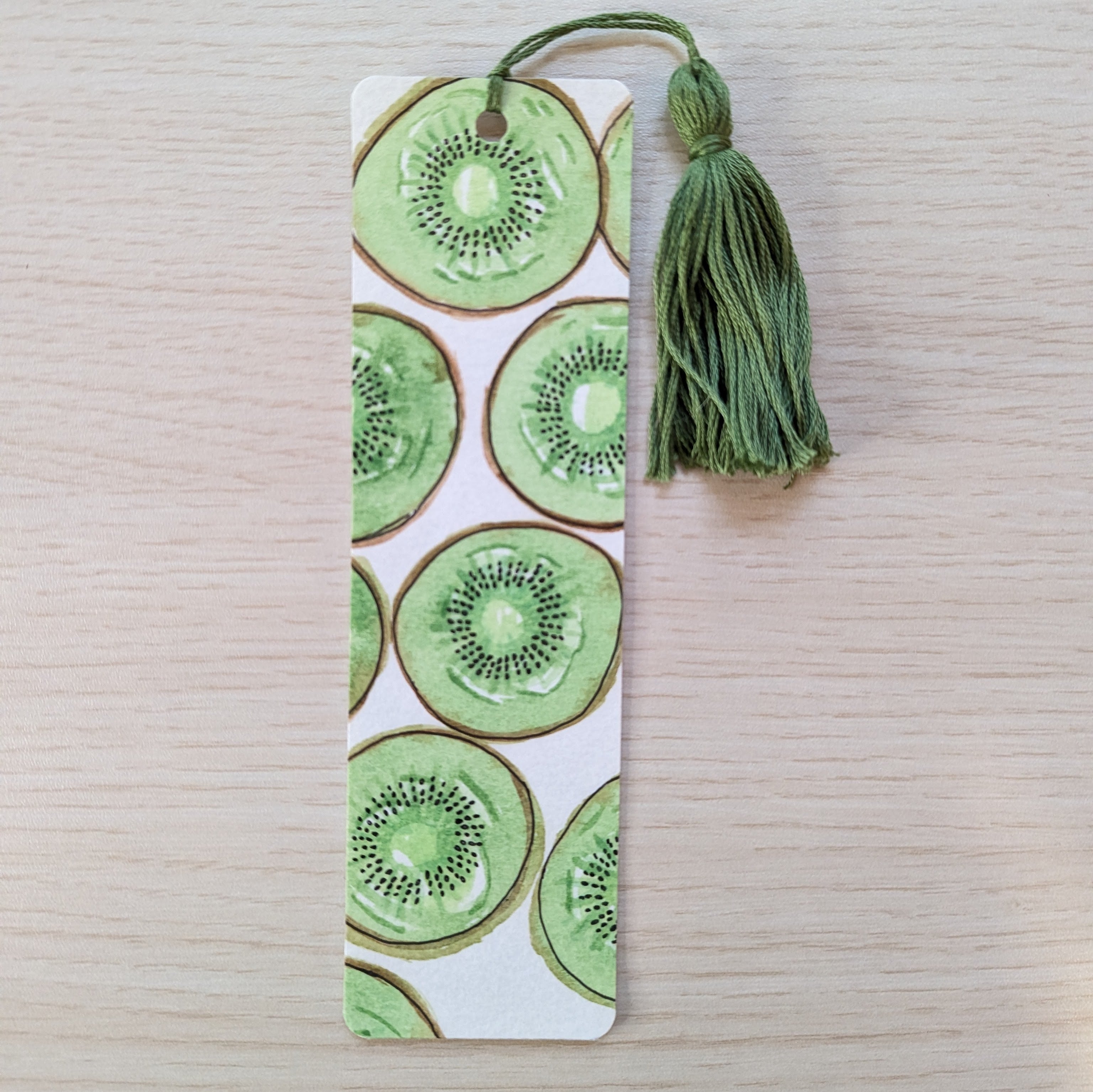 A rectangular paper bookmark with a green kiwi slice pattern and a matching green tassel, placed on a light wood surface.