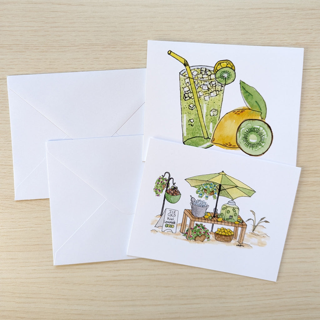 Two illustrated greeting cards with white envelopes on a light wood surface. One card shows a green drink with a straw, lime, and kiwi, and the other shows a small outdoor lemonade stand with an umbrella on a sandy beach.