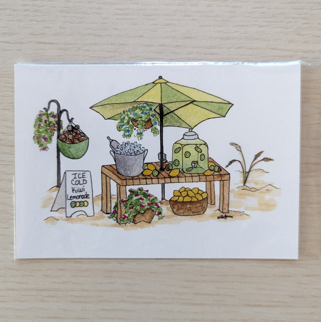 A  white art print featuring a hand-drawn illustration of a lemonade stand under a yellow and green umbrella, placed on a light wood surface.