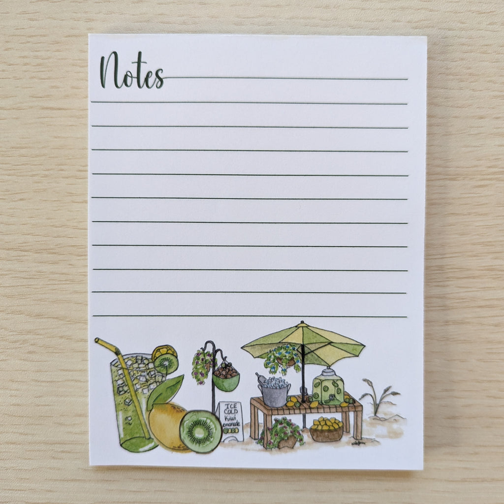 A white lined 'Notes' notepad with a watercolor illustration of a fruit and lemonade stand scene at the bottom, photographed on a light wood surface.