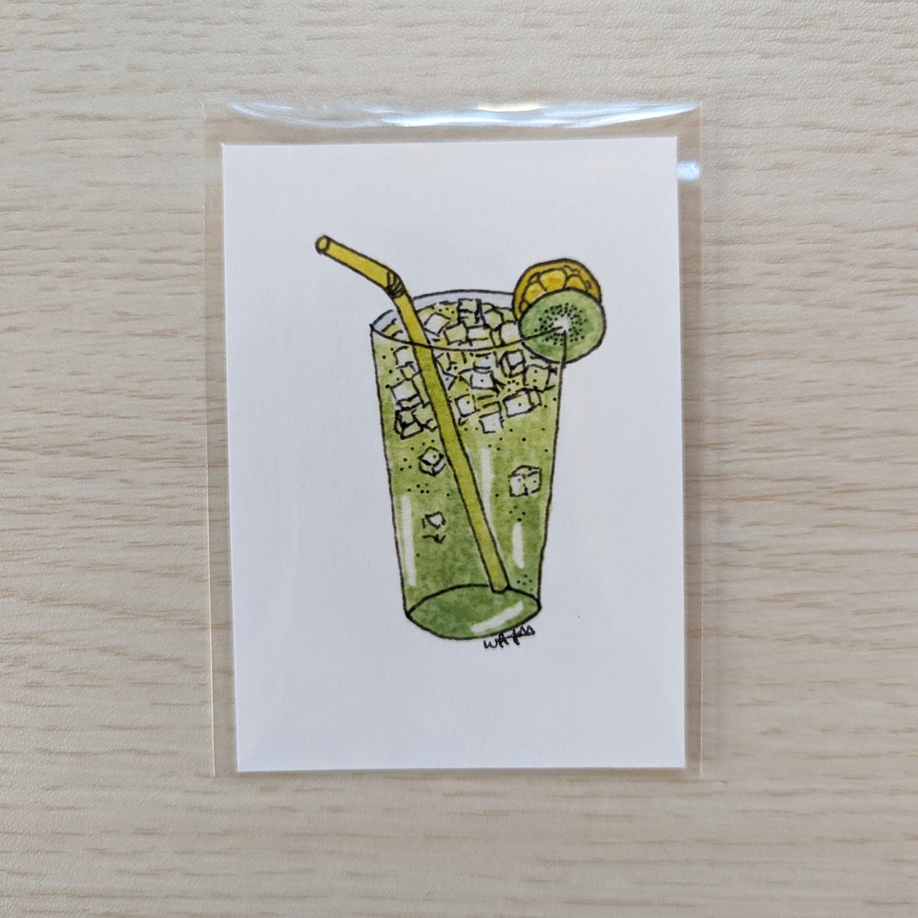 A small white art print featuring a hand-drawn illustration of a green iced drink with a yellow straw and a kiwi slice garnish, photographed on a light wood surface.