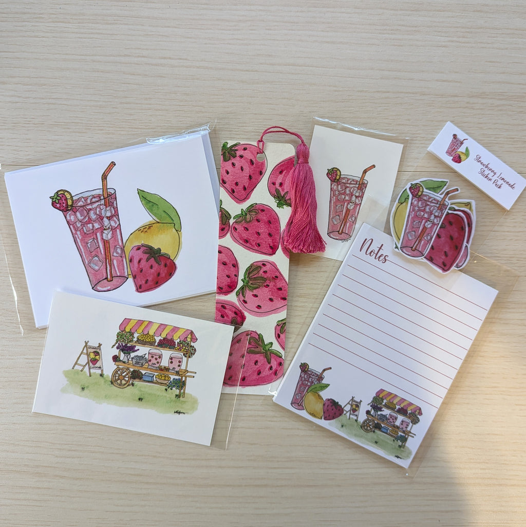A set of strawberry lemonade themed stationery items arranged on a light wood surface, including greeting cards, a lined note pad, a hand painted bookmark with decorative tassel, a sticker pack, and two small illustrated art prints. The illustrations feature pink strawberries, a strawberry lemonade drink, and a lemonade cart.