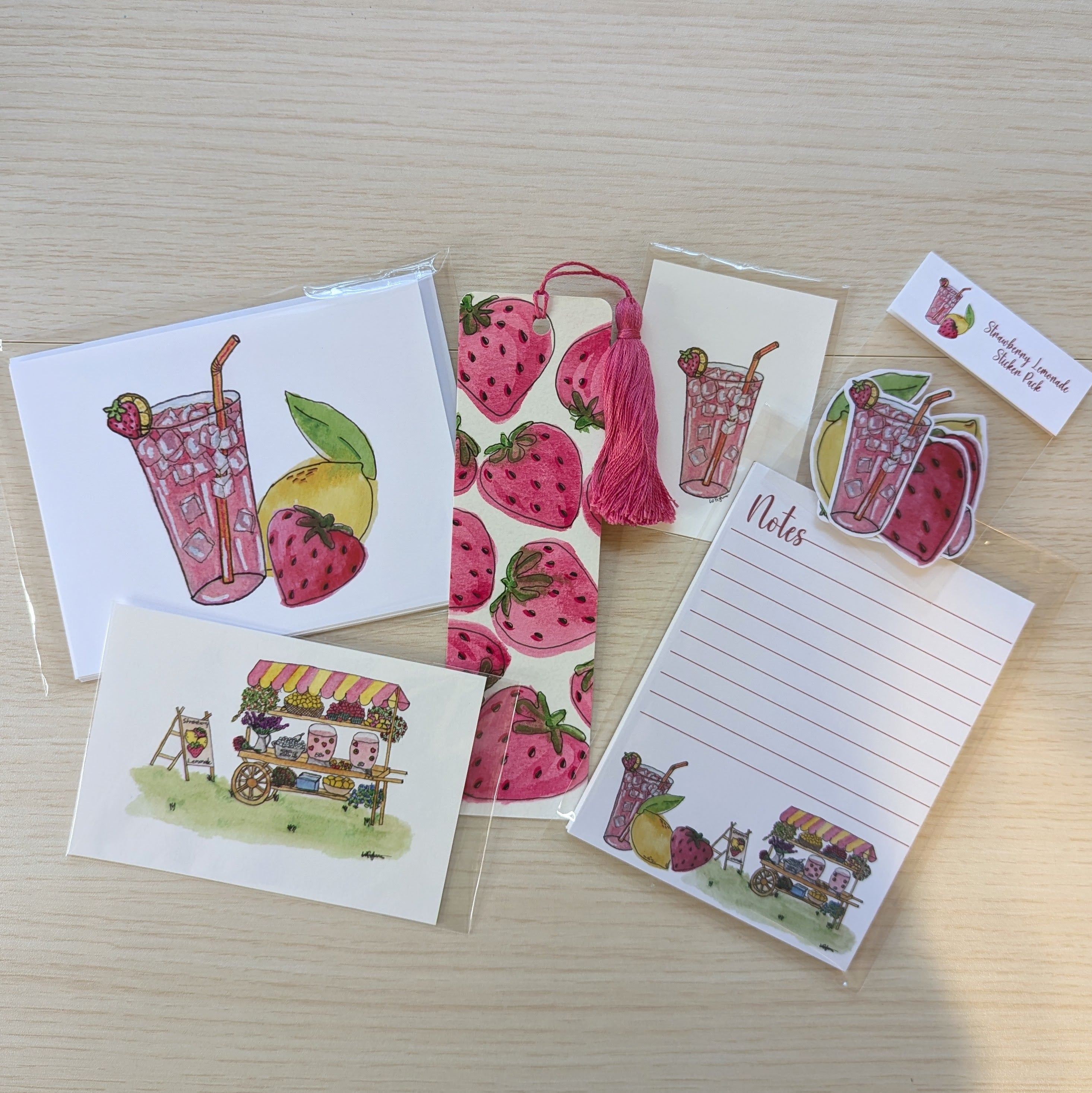 A set of strawberry lemonade themed stationery items arranged on a light wood surface, including greeting cards, a lined note pad, a hand painted bookmark with decorative tassel, a sticker pack, and two small illustrated art prints. The illustrations feature pink strawberries, a strawberry lemonade drink, and a lemonade cart.