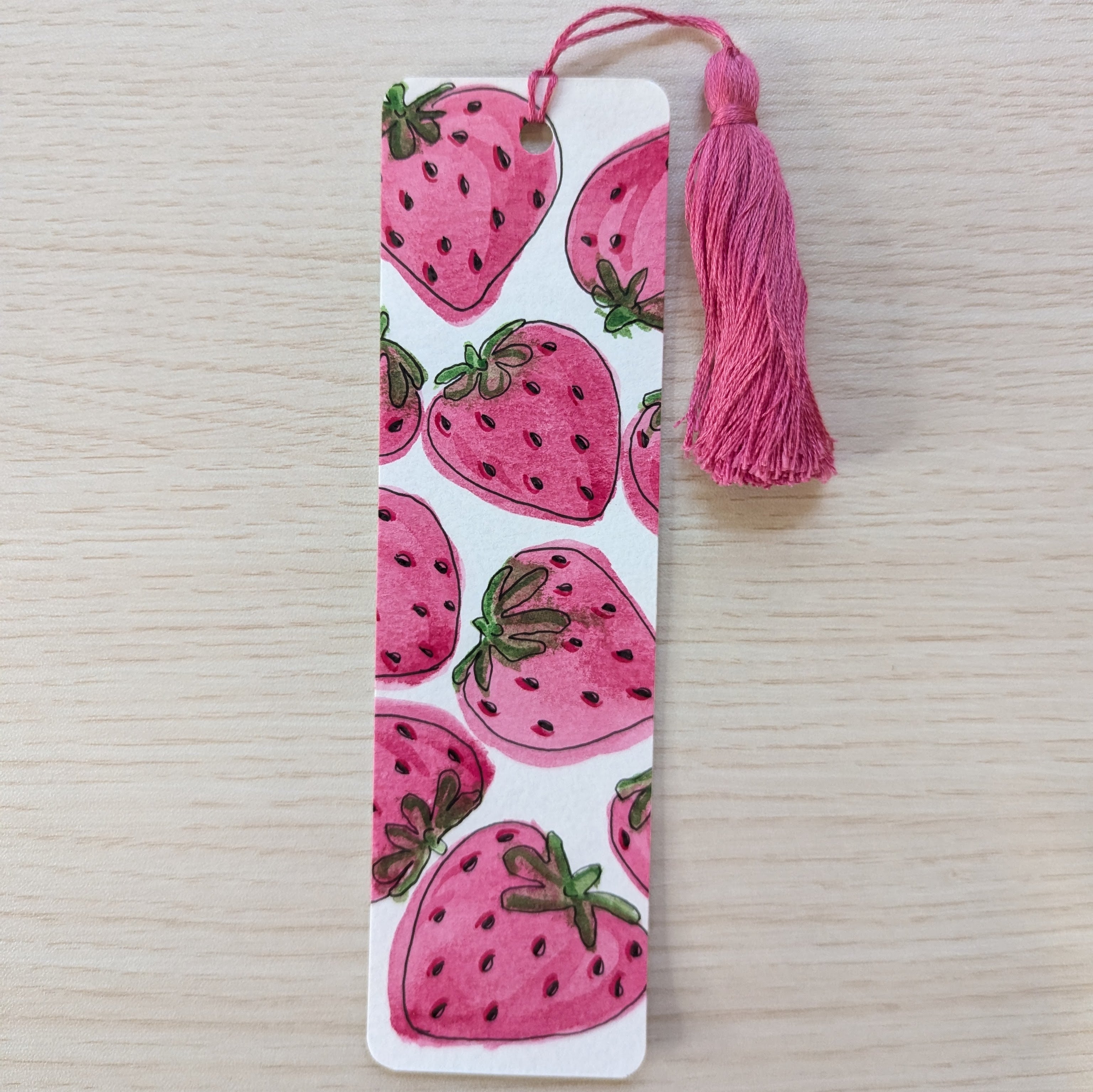 A rectangular paper bookmark with a pink strawberry pattern and a matching pink tassel, placed on a light wood surface.