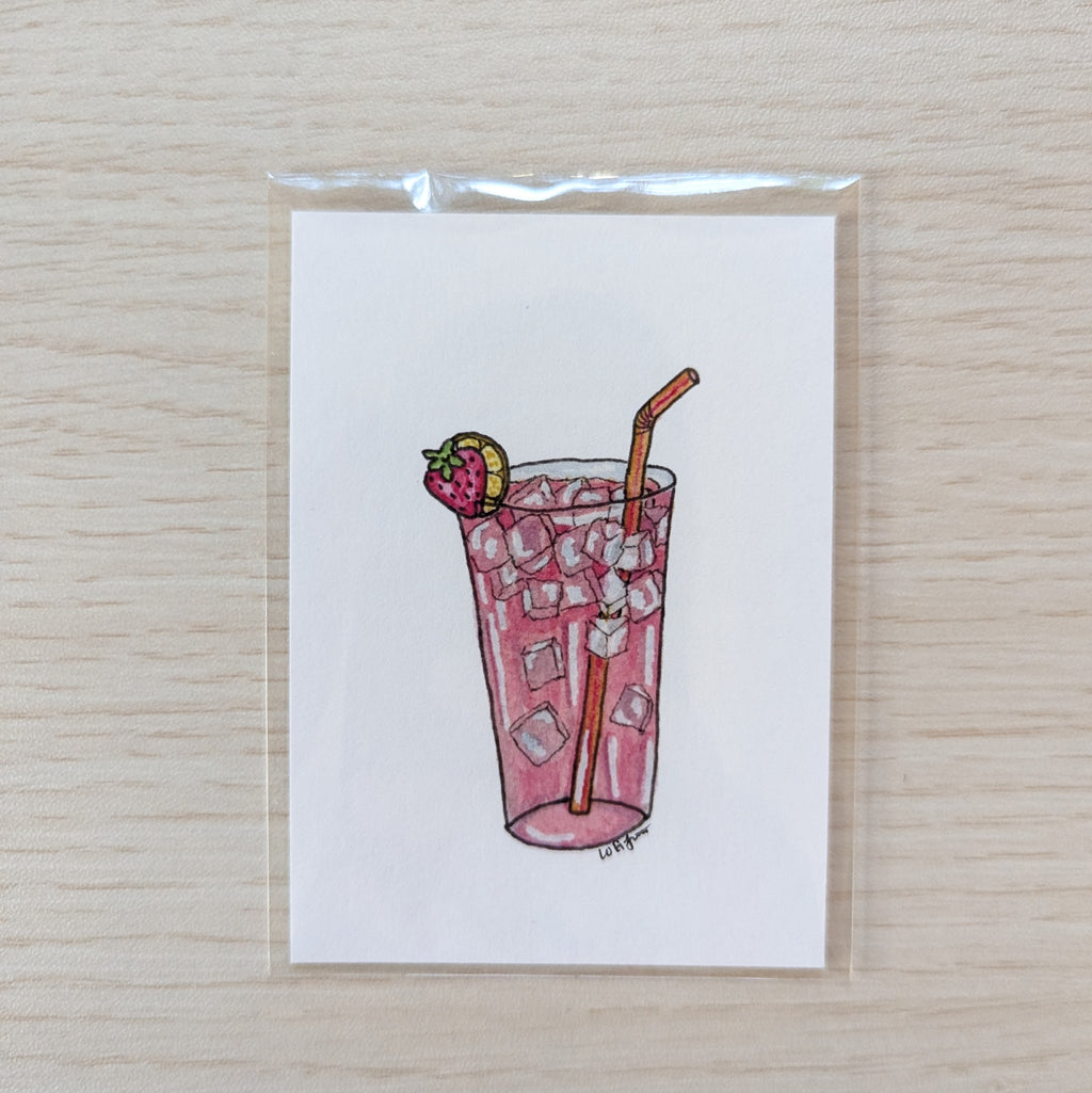 A small illustrated art print of a pink iced drink with a strawberry garnish and a straw, photographed on a light wood surface.