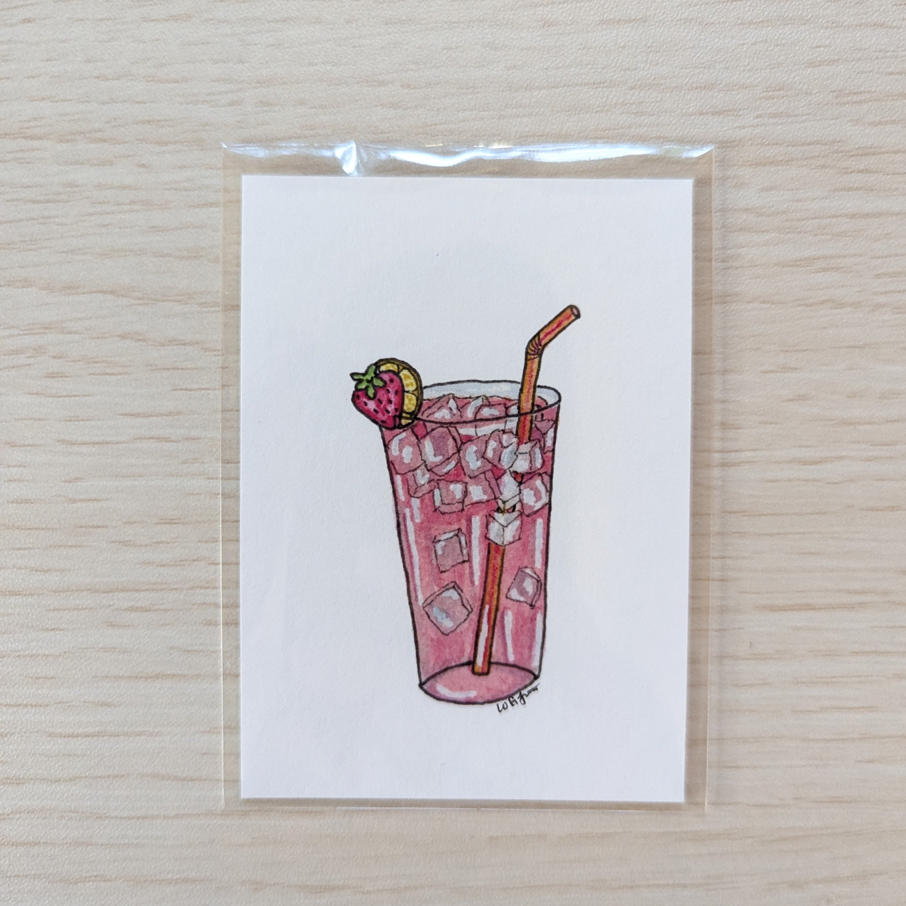 A small illustrated art print of a pink iced drink with a strawberry garnish and a straw, photographed on a light wood surface.