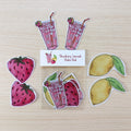 A sticker pack featuring strawberry, lemon, and pink drink illustrations arranged on a light wood background, with a small label reading 'Strawberry Lemonade Sticker Pack'.