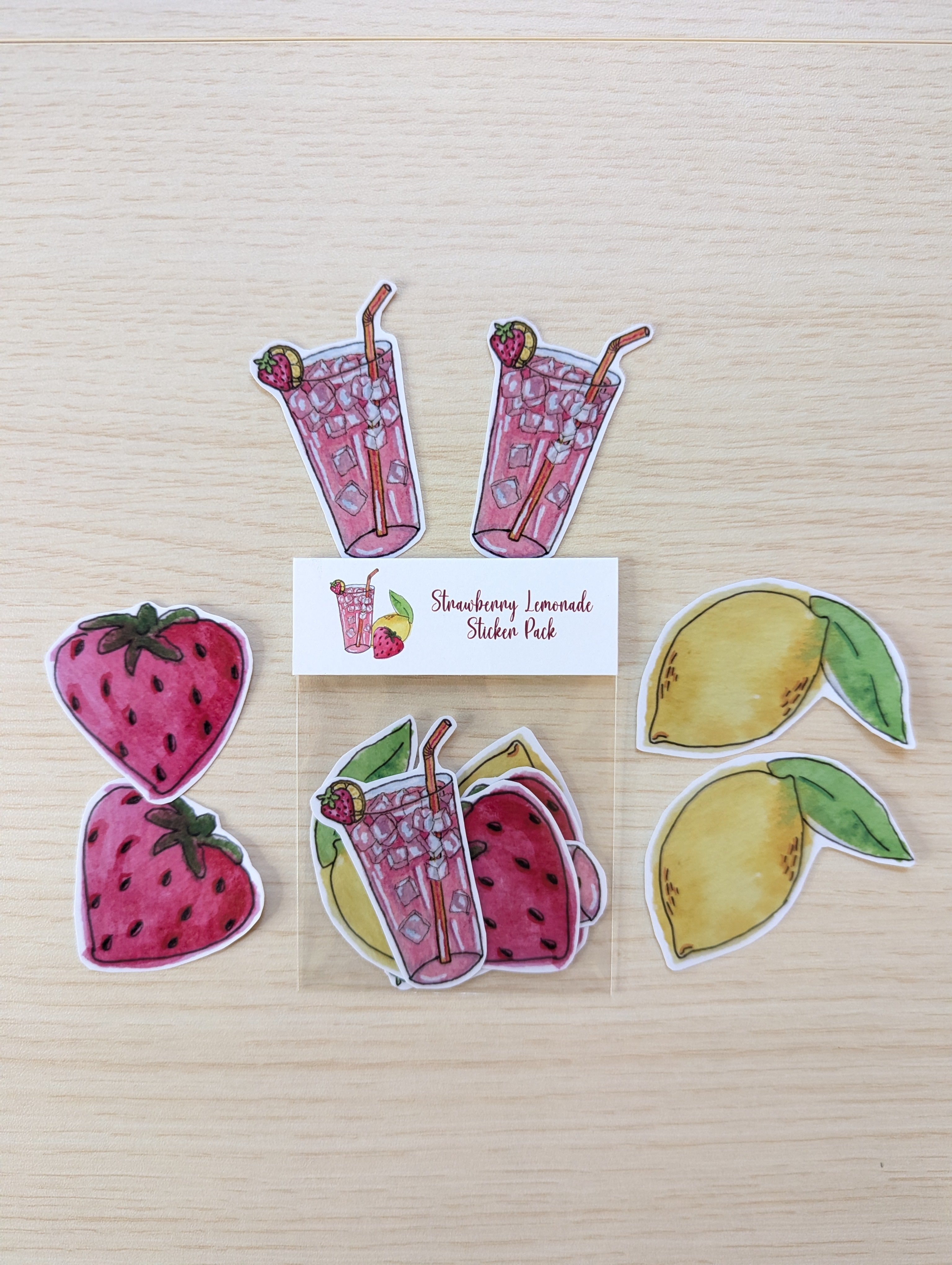 A sticker pack featuring strawberry, lemon, and pink drink illustrations arranged on a light wood background, with a small label reading 'Strawberry Lemonade Sticker Pack'.