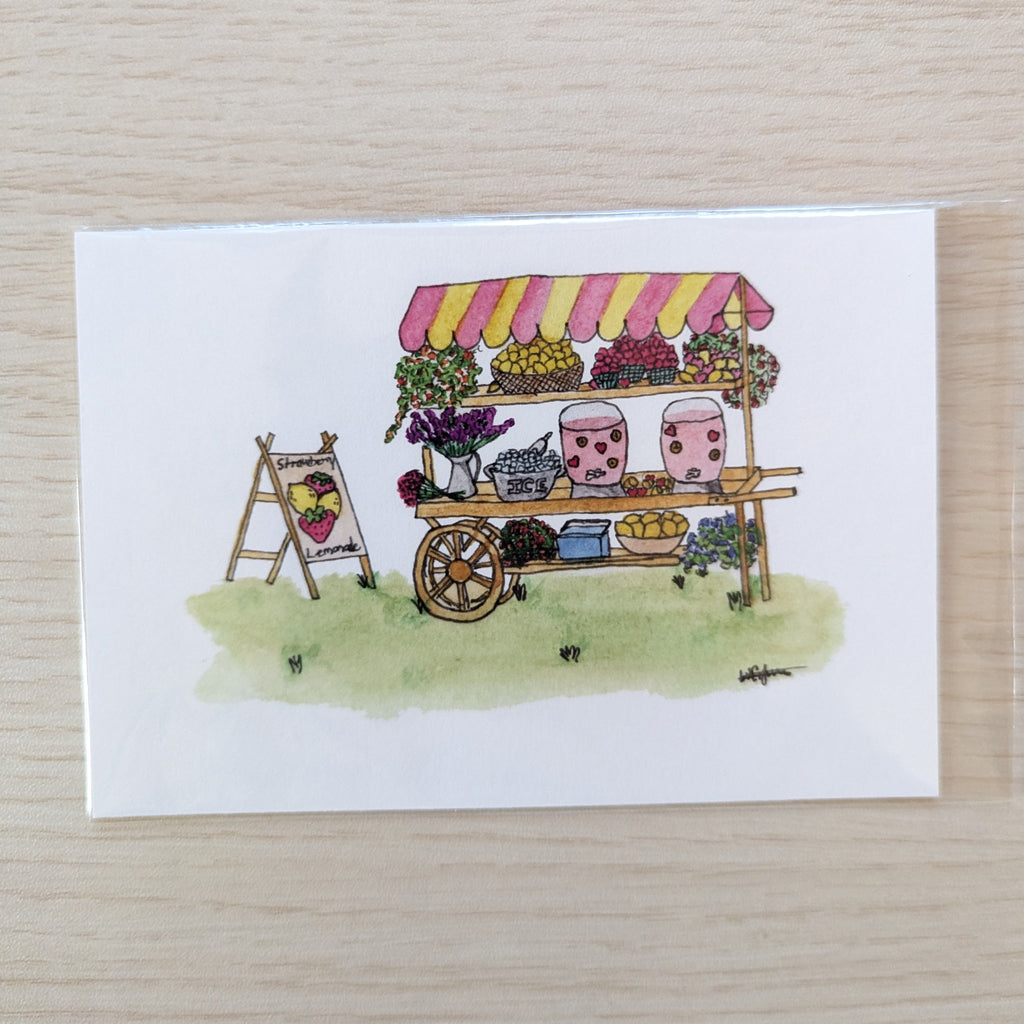 A white art print featuring a hand-drawn illustration of a small market cart with a pink and yellow striped awning, flowers, jars, and fruit, placed on a light wood surface.