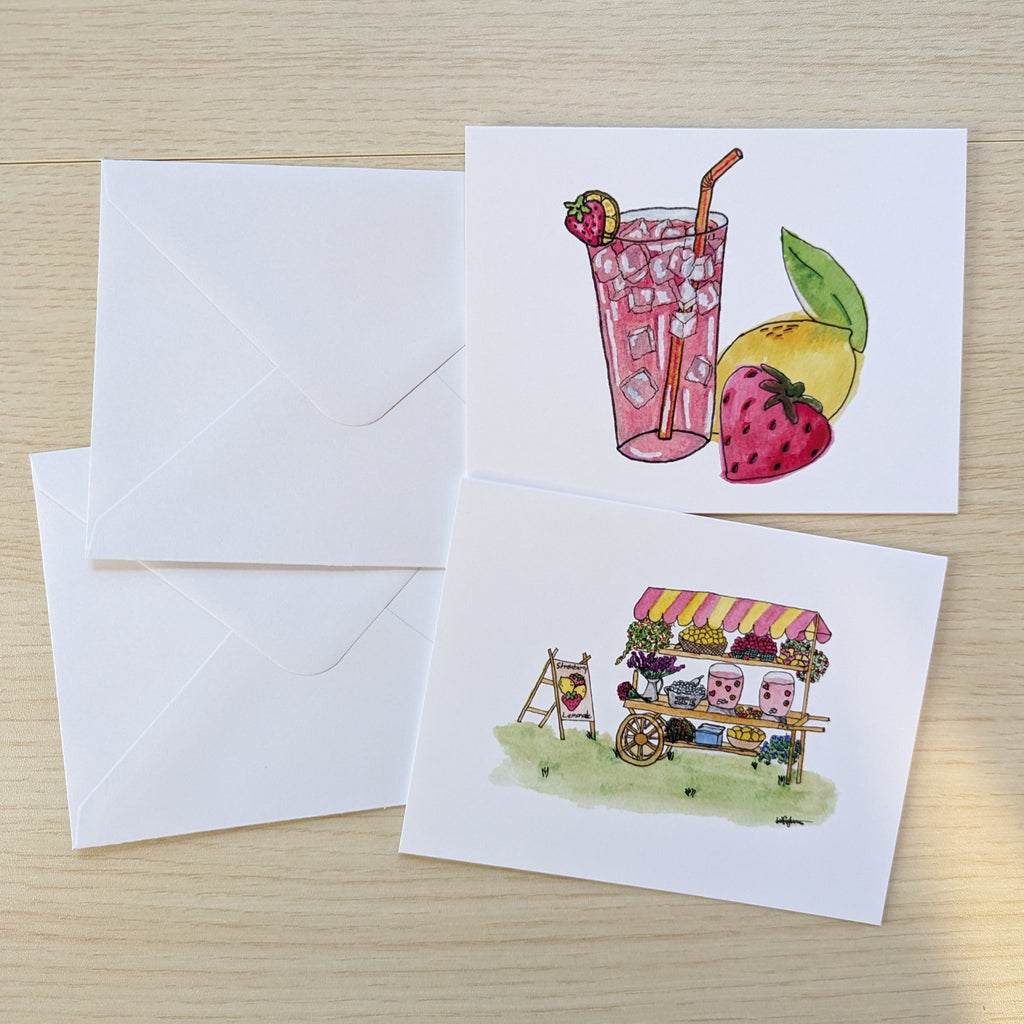 Two white greeting cards with watercolor illustrations and two white envelopes on a light wood surface. The top card shows a pink drink with a strawberry garnish and a yellow fruit, and the bottom card shows a small market stall with flowers and pink lemonade.