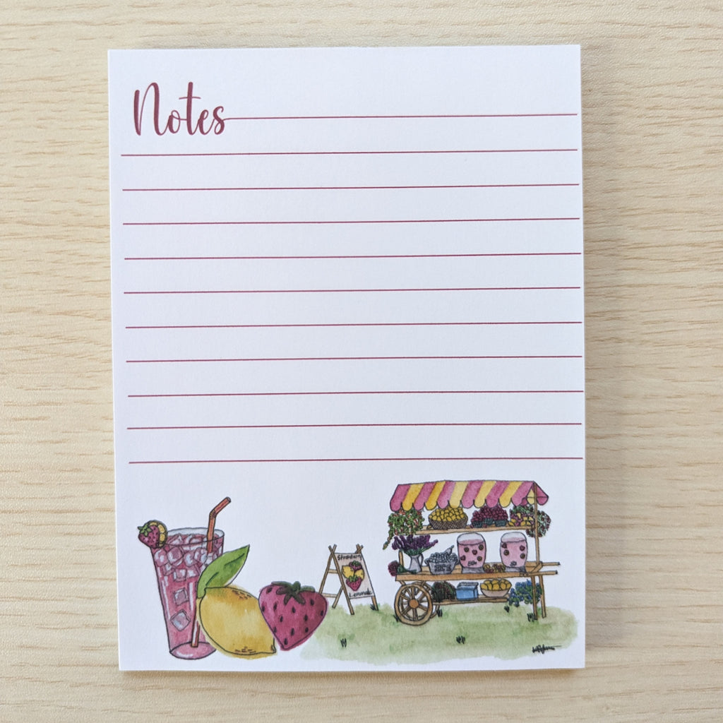 A white lined notepad with the word 'Notes' at the top left and a small watercolor illustration along the bottom depicting a drink, a lemon, a strawberry, and a market cart, placed on a light wood surface.