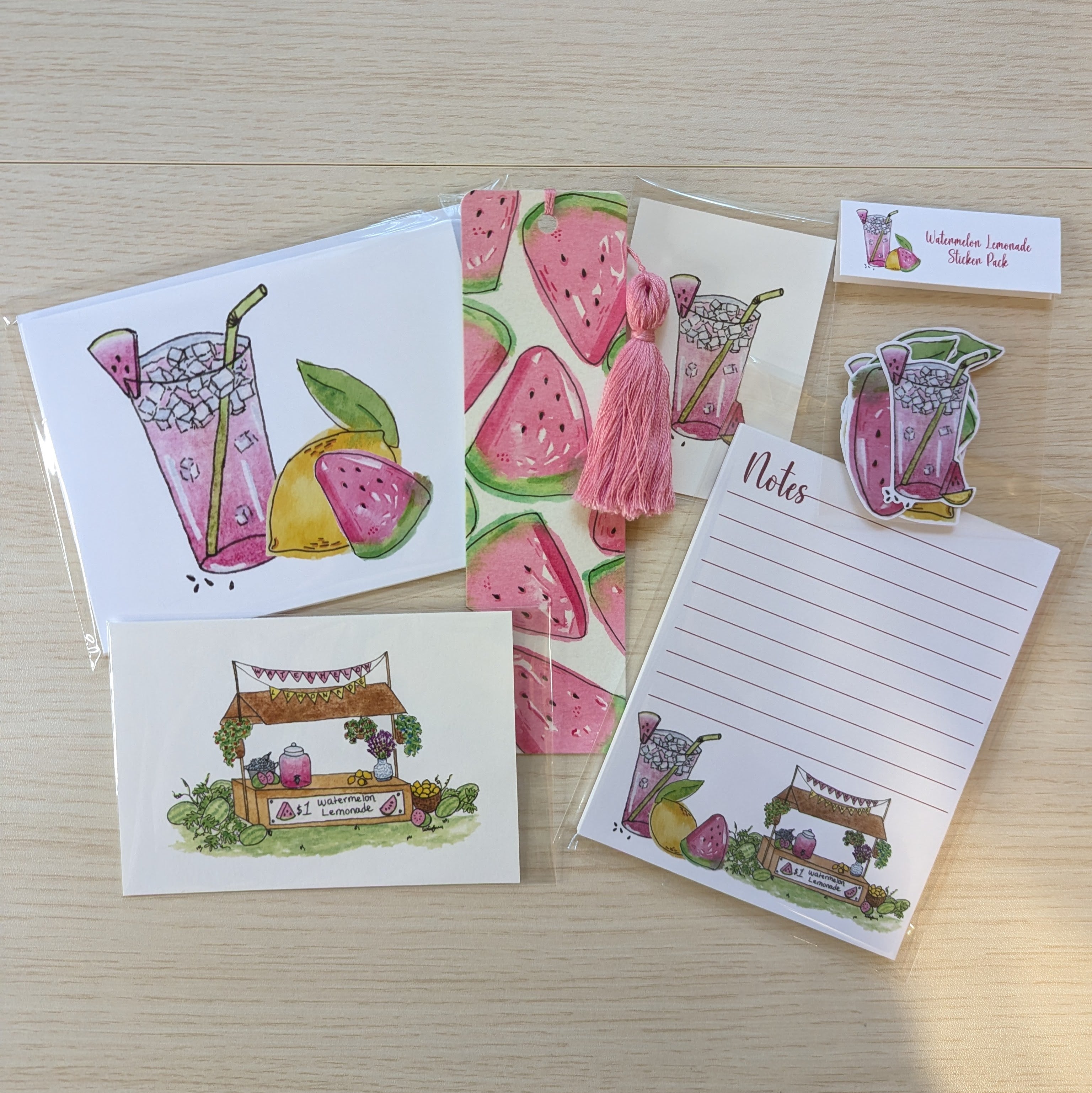 A set of watermelon-themed stationery items arranged on a light wood surface, including illustrated cards, a hand painted watermelon bookmark and pink tassel, a notepad, stickers, and two small illustrated art prints.