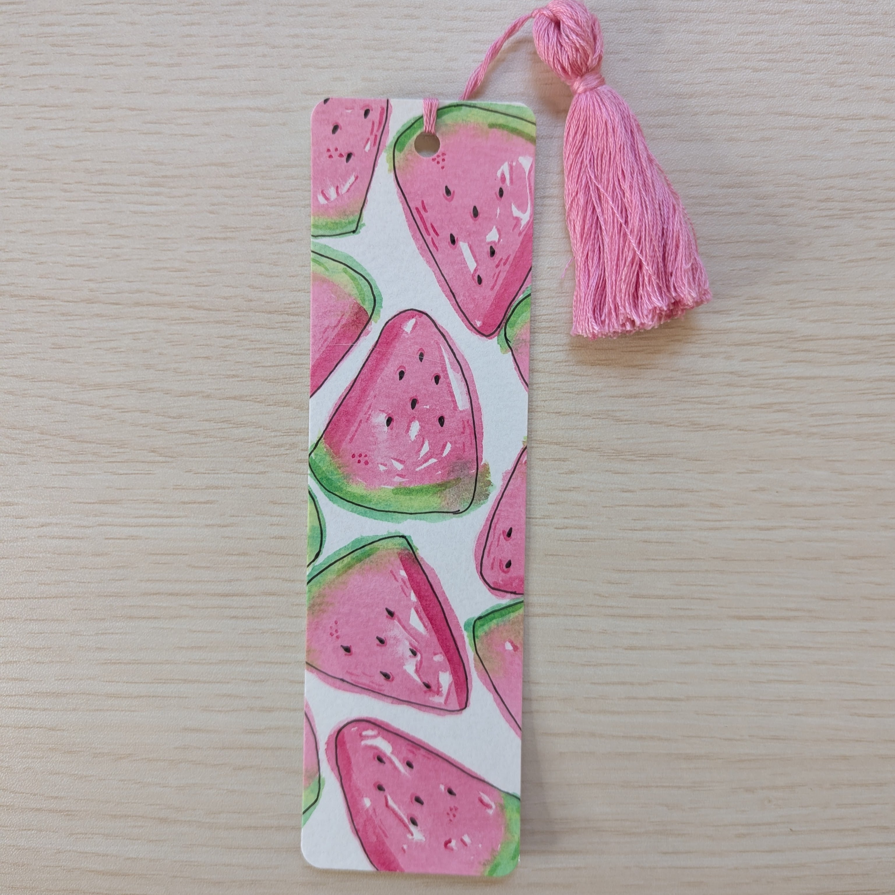 A rectangular paper bookmark with a pink watermelon slice pattern and a matching pink tassel, placed on a light wood surface.