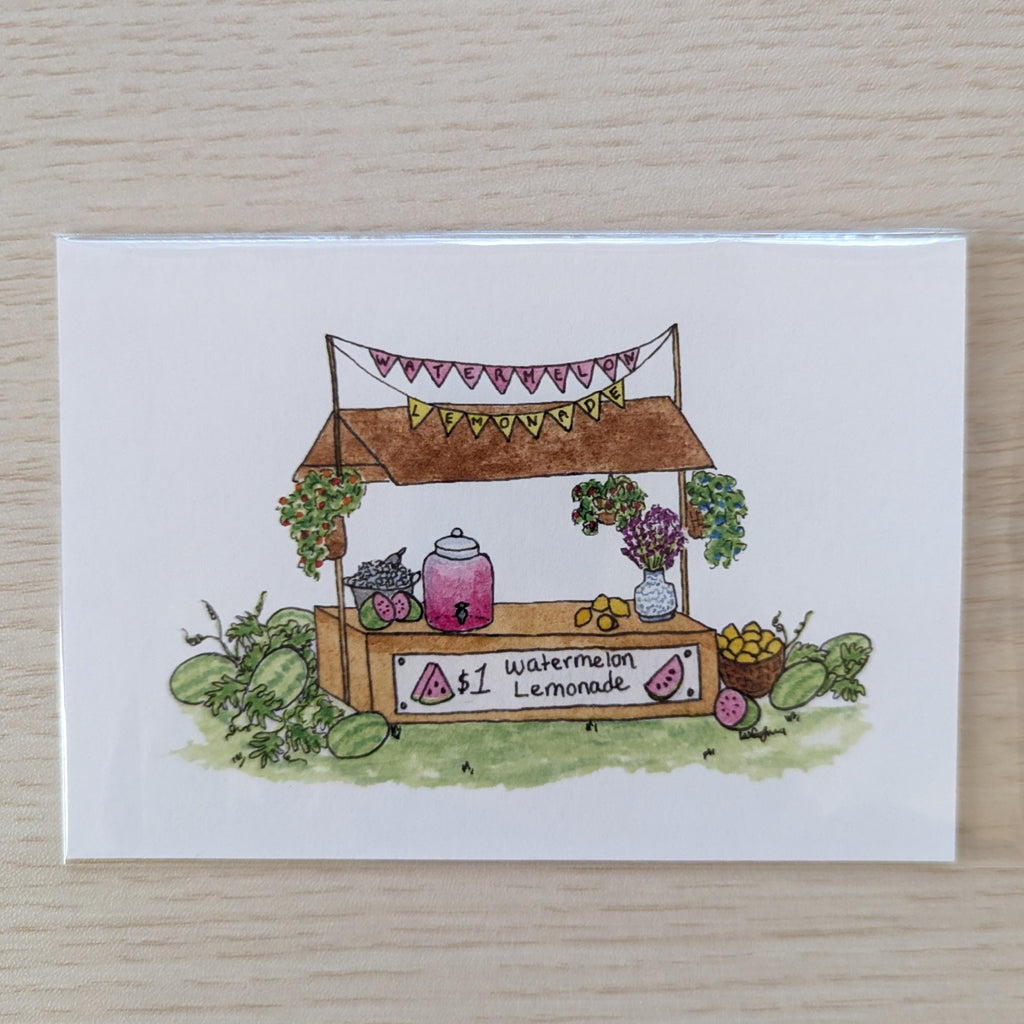 A white art print featuring a hand-drawn watercolor illustration of a lemonade stand with watermelons, flowers, and a sign reading '$1 Watermelon Lemonade', placed on a light wood surface.