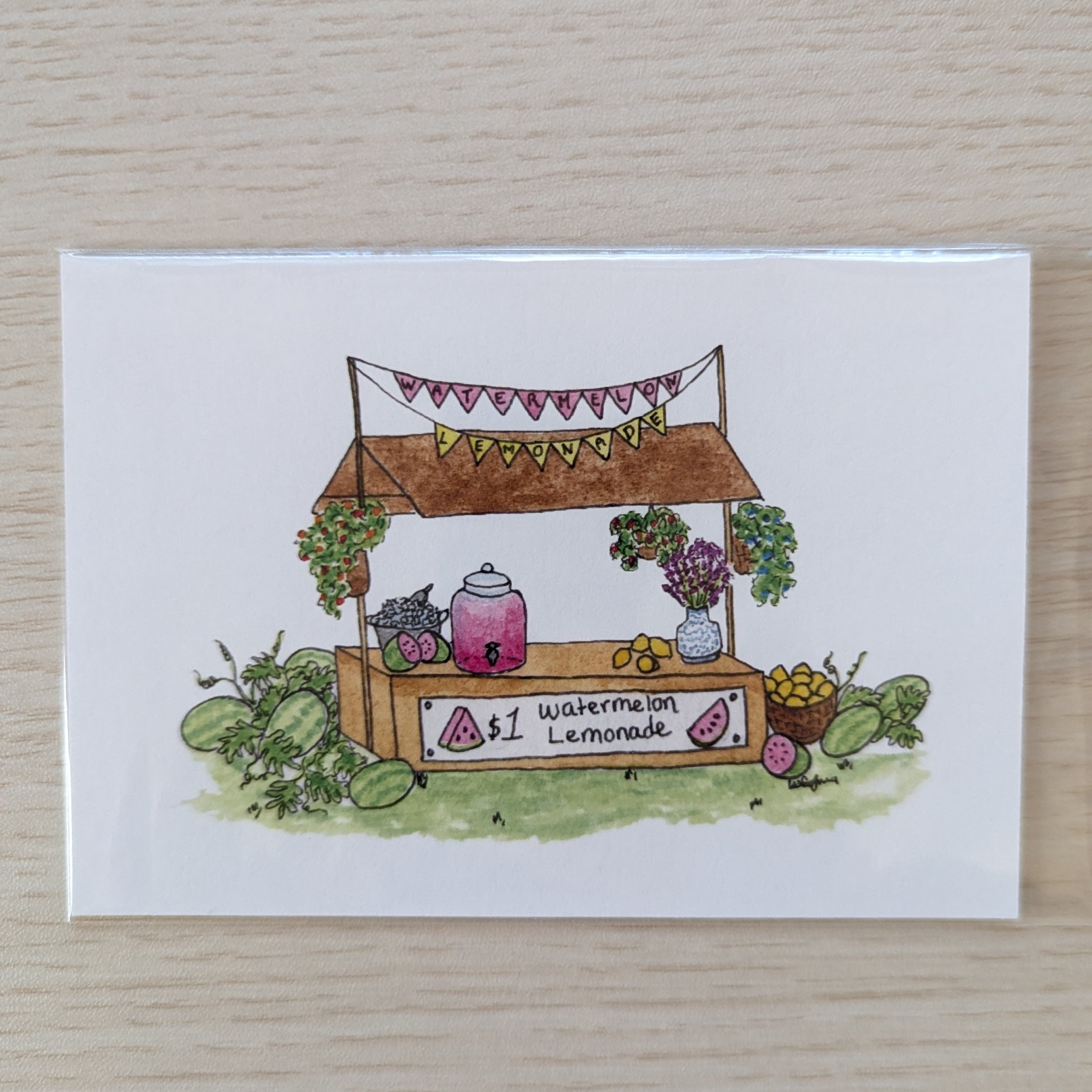 A white art print featuring a hand-drawn watercolor illustration of a lemonade stand with watermelons, flowers, and a sign reading '$1 Watermelon Lemonade', placed on a light wood surface.