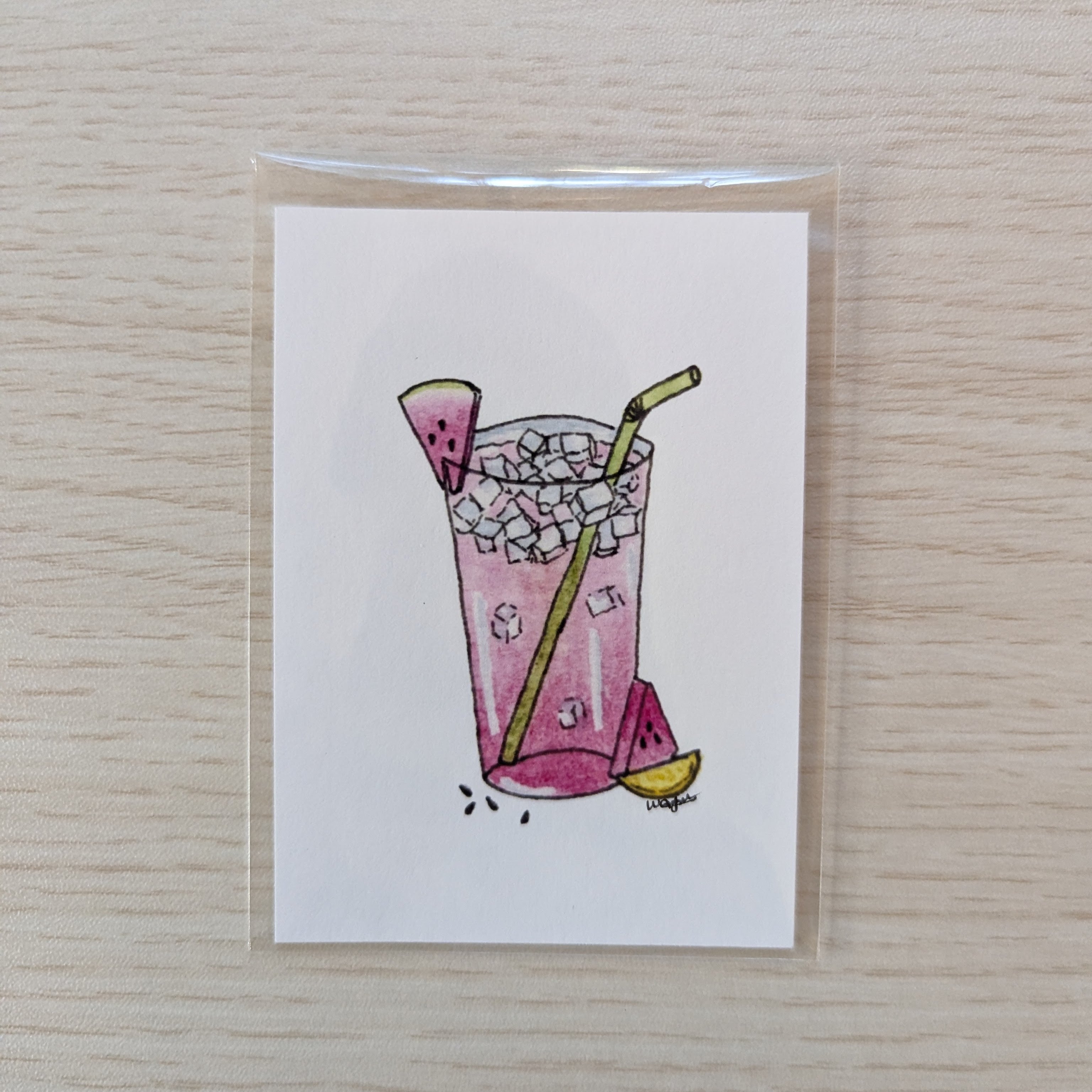 A white art print featuring a hand-drawn pink iced drink with watermelon and lemon slices, photographed on a light wood surface.