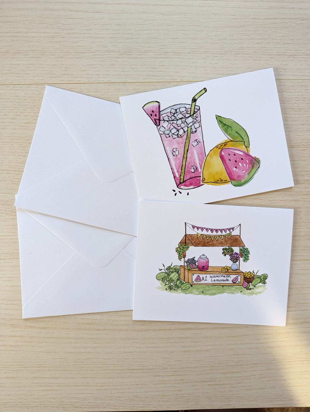 Two white greeting cards with watercolor illustrations on a light wood surface, alongside two white envelopes. The top card shows a pink drink with watermelon and lemon graphics, and the bottom card shows a small lemonade stand with fruit and flowers.