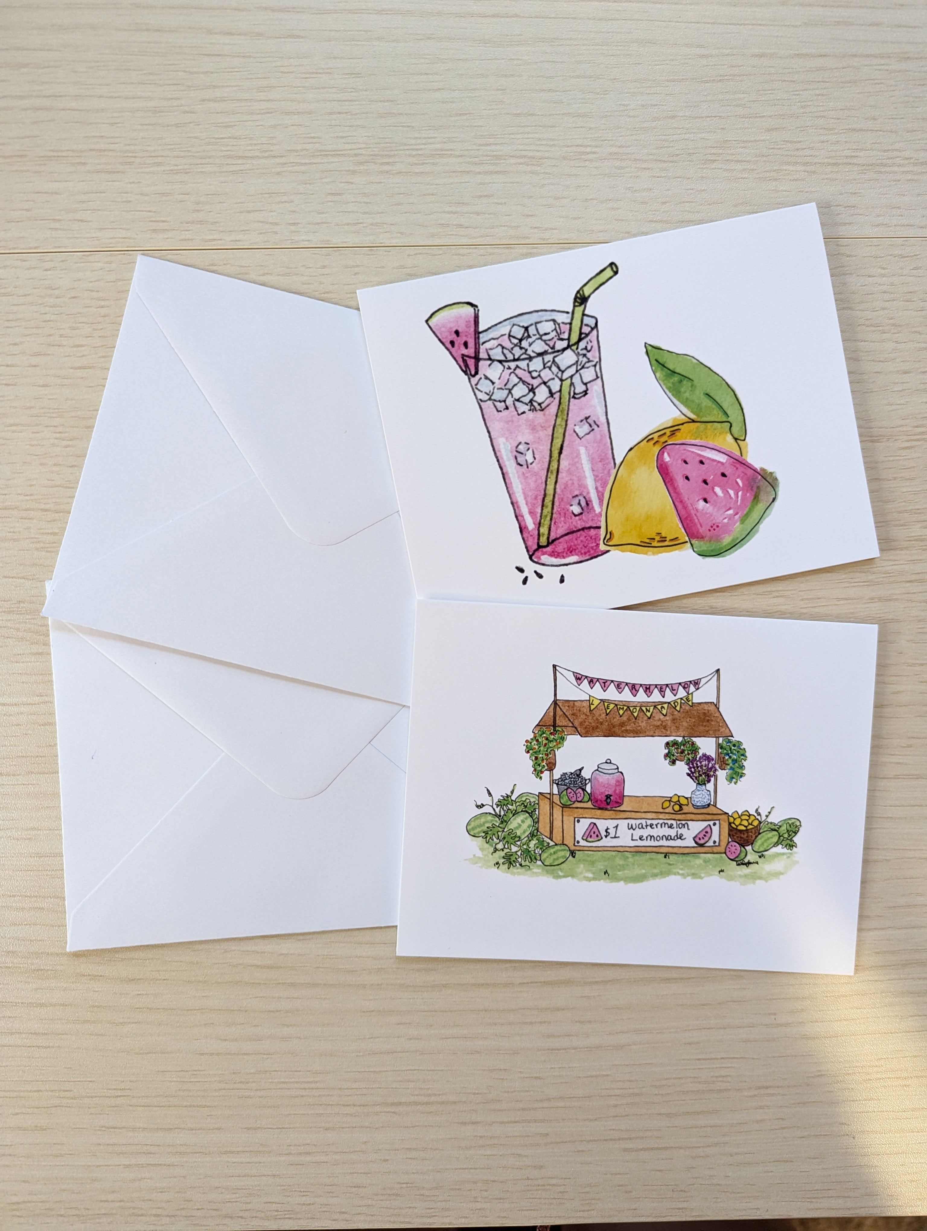 Two white greeting cards with watercolor illustrations on a light wood surface, alongside two white envelopes. The top card shows a pink drink with watermelon and lemon graphics, and the bottom card shows a small lemonade stand with fruit and flowers.