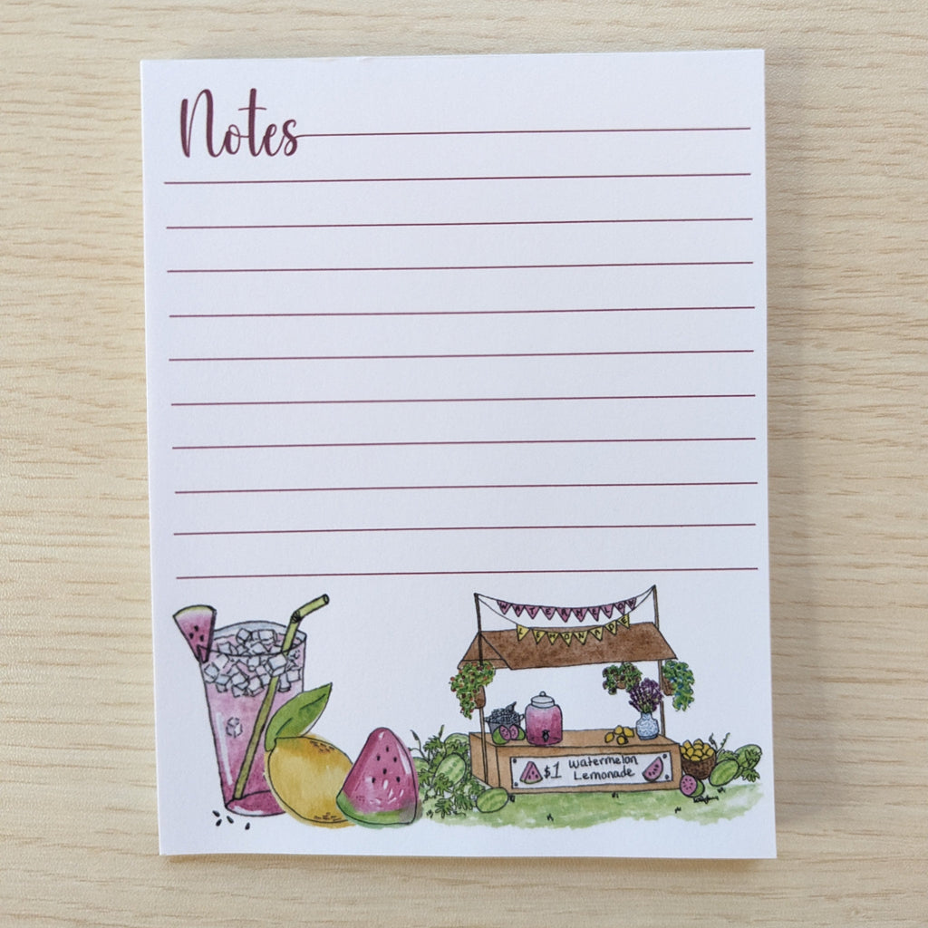 A small lined notepad with the word 'Notes' at the top left and a watercolor illustration along the bottom showing a drink, fruit, and a market stall, placed on a light wood surface.