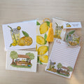 A set of lemon-themed stationery items including a notepad, greeting cards, a tassel bookmark, and stickers, all featuring illustrations of lemons, iced drinks, and a lemonade stand, arranged on a light wood background.