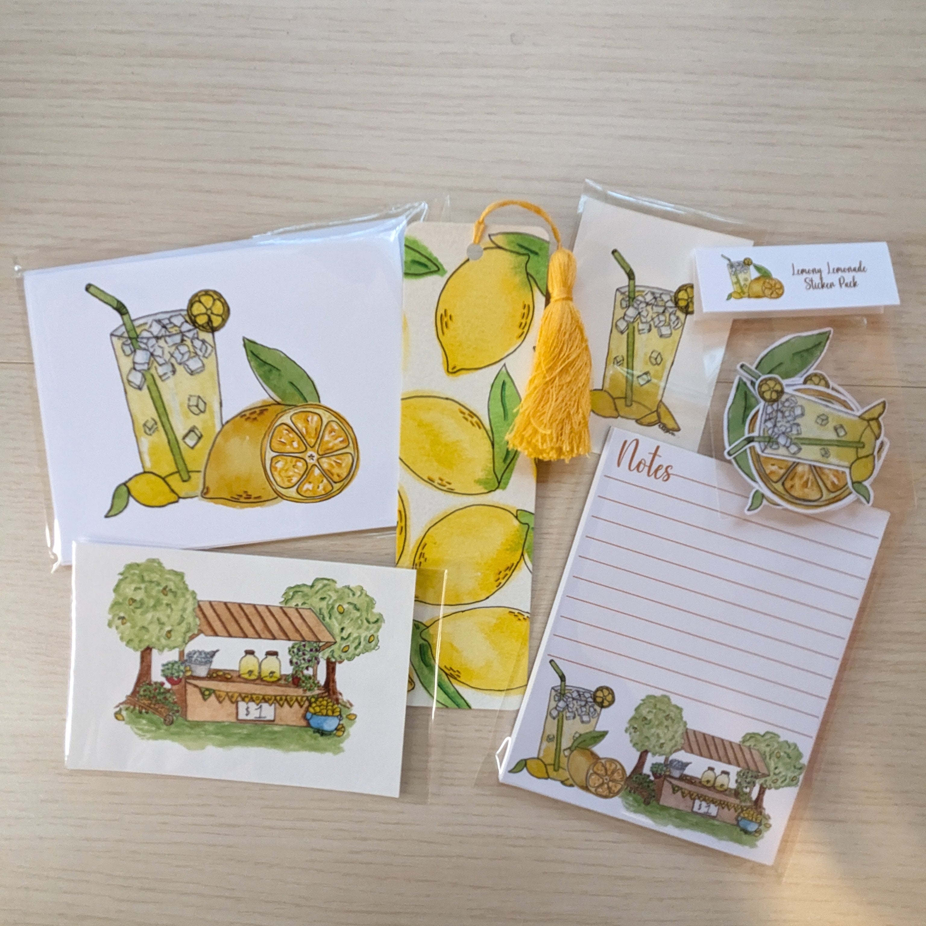 A set of lemon-themed stationery items including a notepad, greeting cards, a tassel bookmark, and stickers, all featuring illustrations of lemons, iced drinks, and a lemonade stand, arranged on a light wood background.