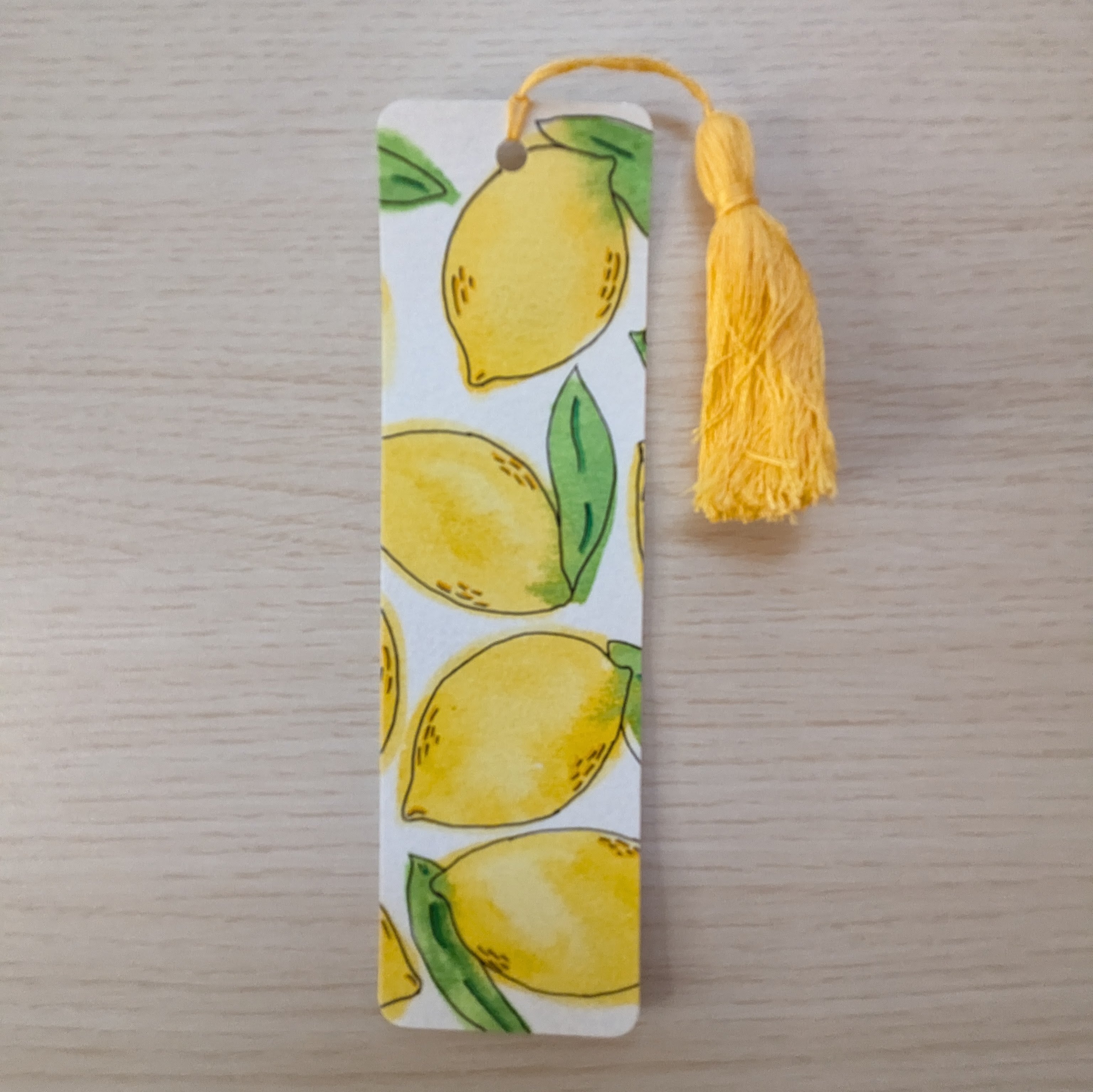 A rectangular paper bookmark with a yellow lemon and green leaf pattern, featuring a yellow tassel at the top, displayed on a light wood surface.