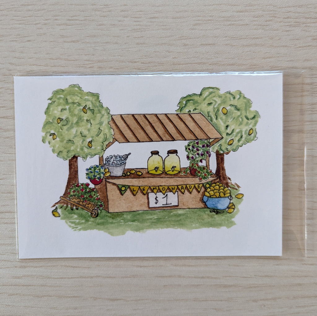 A square white art print featuring a hand-drawn watercolor illustration of a small lemonade stand with two leafy lemon trees, jars of lemonade, flowers, and a basket, placed on a light wood surface.