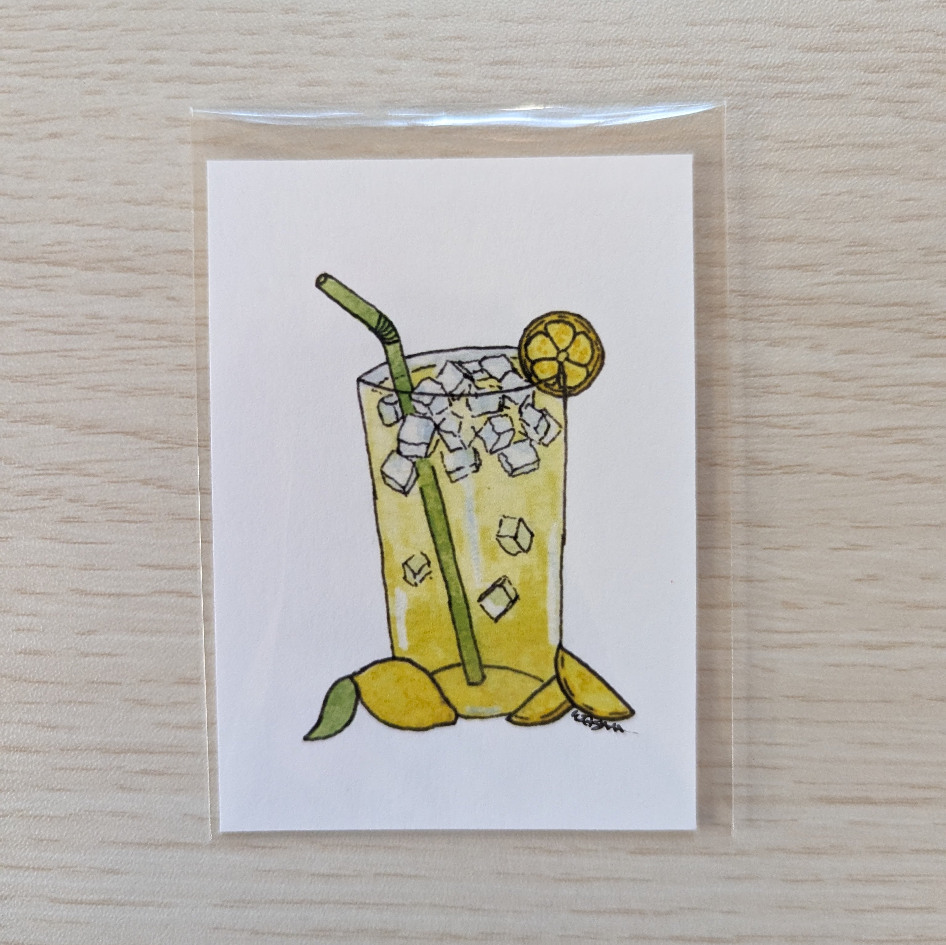 A square white ACEO size art print featuring a hand-drawn illustration of a lemonade glass with ice, a green straw, yellow lemon slices, and a green leaf, photographed on a light wood surface.