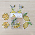 A set of illustrated lemon-themed sticker cutouts on a light wood surface, including lemon slices, whole lemons, and two tall lemonade glasses with straws. A small card in the center reads "Lemony Lemonade Sticker Pack.
