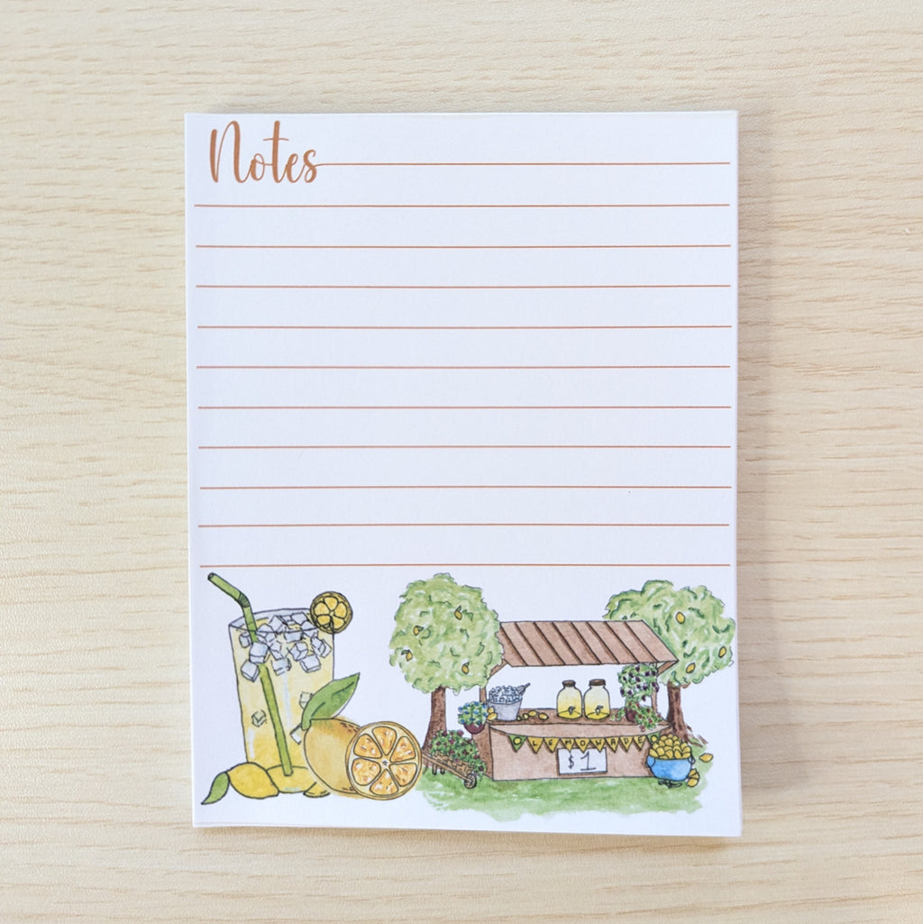 A lined notepad with the word 'Notes' at the top and a small illustrated lemonade stand and drink at the bottom, photographed on a light wood surface.