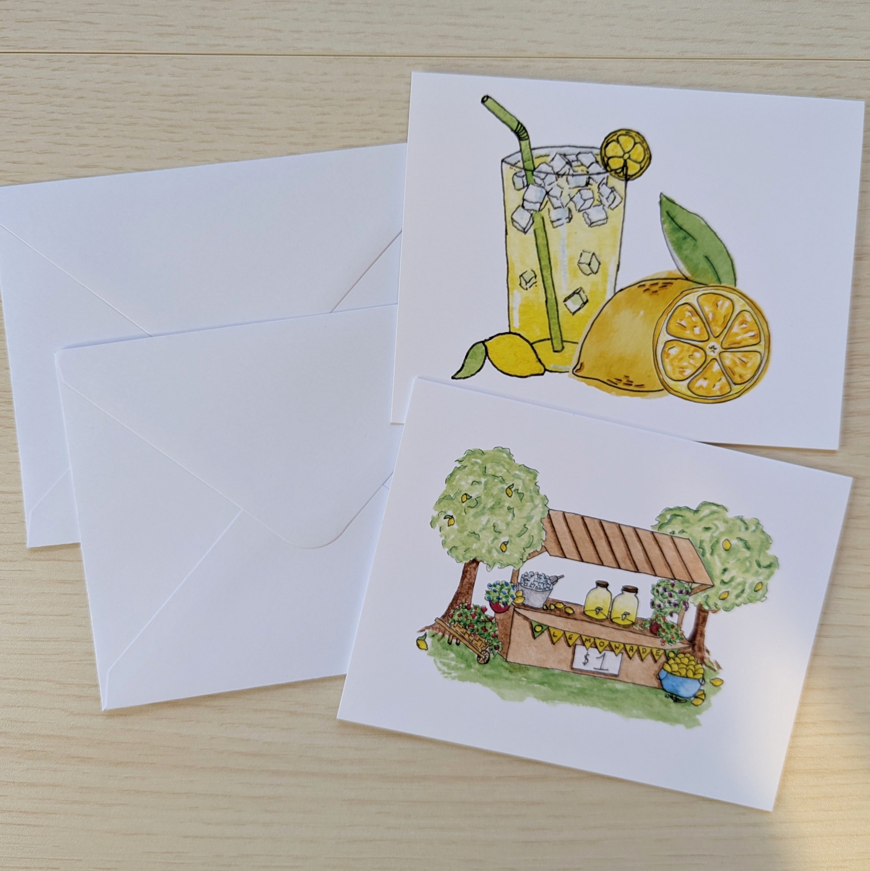 Two white greeting cards with hand-drawn lemon-themed illustrations and matching white envelopes, arranged on a light wood surface.